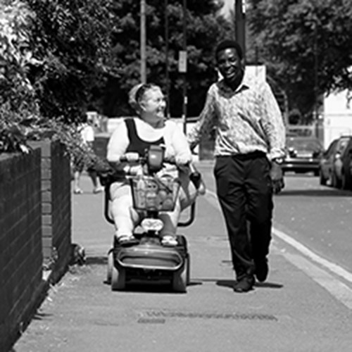 Woman in a mobility scooter going down a pavement with a personal assistant carer