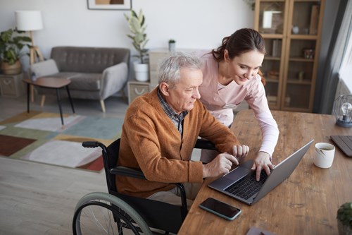 Man over 50 in a wheel chair being helped by a younger woman with his laptop