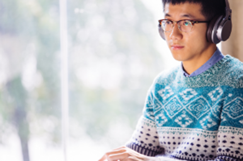 Young man wearing glasses and headphones and a patterned blue jumper working by a window 