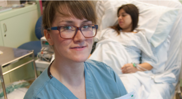 Midwife wearing glasses looking towards the camera with a pregnant women in the background 