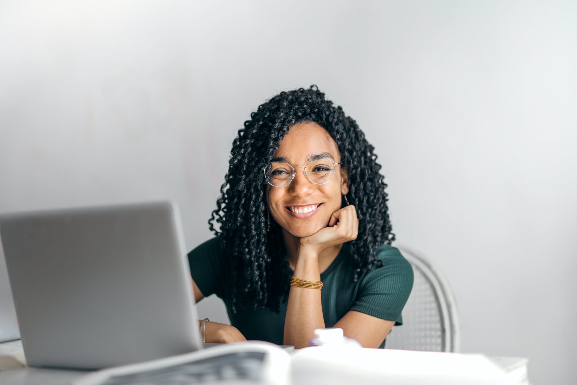 Woman with black curly hair and glasses smiling at the camera using a laptop 