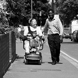 Woman in a mobility scooter going down a pavement with a personal assistant carer